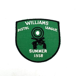 May include: A shield-shaped green embroidered patch with a black border. It features the white text WILLIAMS PISTOL LEAGUE, a black target icon, two crossed black pistols, and the white text SUMMER 1958 centered on the fabric surface.