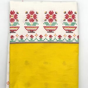 May include: Folded white and yellow textile with embroidered floral patterns. The top section features red, yellow, and green cross-stitch flowers. The bottom half is solid yellow. The textile is folded, showing the design.