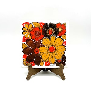May include: A square ceramic tile featuring a vibrant floral pattern in shades of orange, yellow, brown, and red. The design includes large, stylized blossoms and leaves. The signature Georges Briard is printed in black in the bottom right corner.