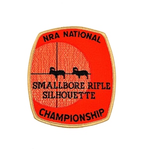 May include: A rectangular embroidered patch with rounded corners and a gold border. The background is bright orange with a black crosshair graphic, two black ram silhouettes, and the black text NRA NATIONAL SMALLBORE RIFLE SILHOUETTE CHAMPIONSHIP.
