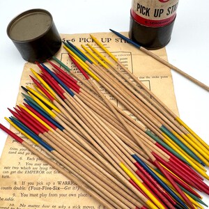 May include: A vintage Pick-Up Sticks game set. The game includes a cylindrical container, a metal tin, and a collection of wooden sticks with colored tips in red, yellow, blue, and green. The instructions are visible on a paper sheet.