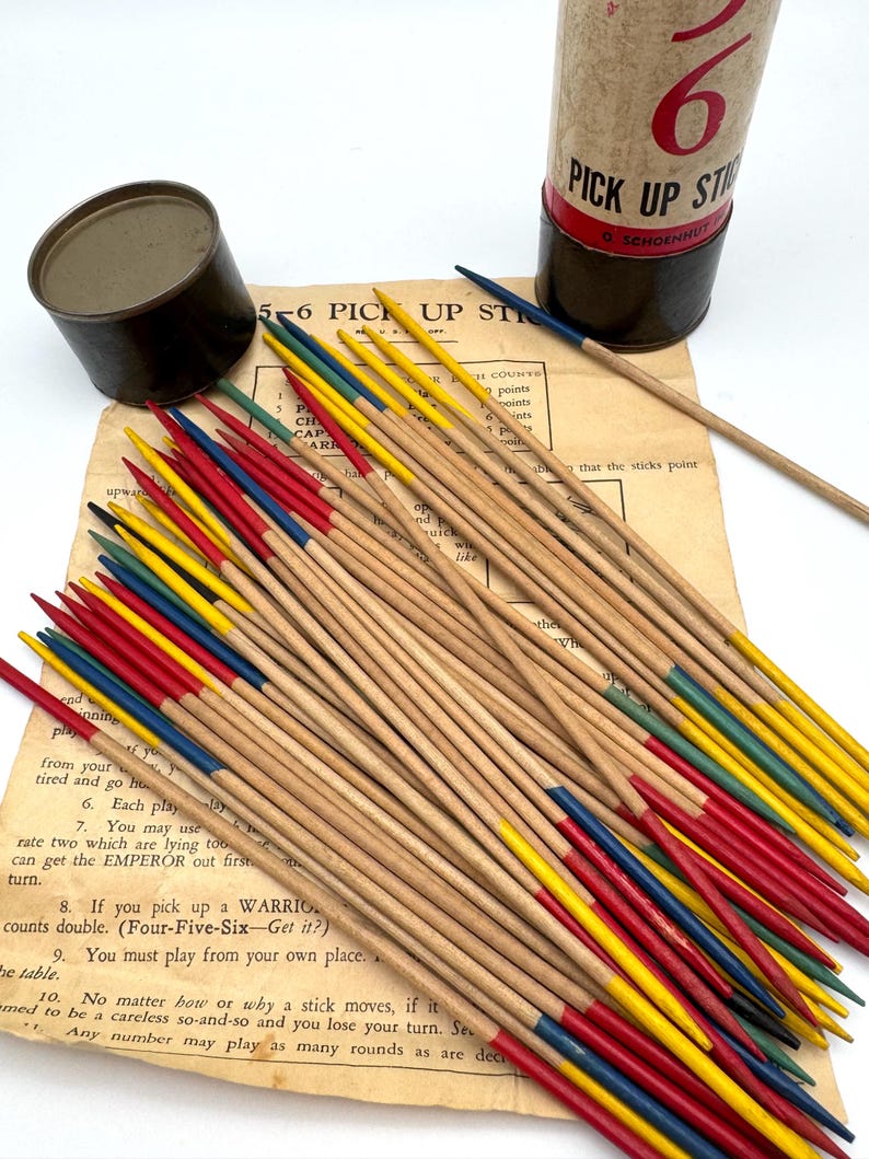 May include: A vintage Pick-Up Sticks game set. The game includes a cylindrical container, a metal tin, and a collection of wooden sticks with colored tips in red, yellow, blue, and green. The instructions are visible on a paper sheet.