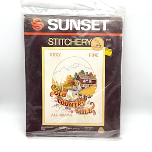 May include: A Sunset Stitchery kit featuring an "Old Country Mill" design. The package is sealed in clear plastic and has a brown border with the Sunset logo. The design depicts a mill with a water wheel, wheat, and the words "Old Country Mill".