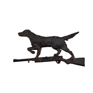 May include: A dark brown, cast metal wall hook rack shaped like a hunting dog standing on top of a rifle. The rifle features four small hooks extending from the bottom edge, designed for hanging items. The metal has a weathered, textured finish.