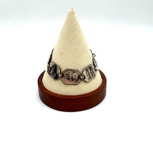 May include: Silver-toned bracelet with rectangular links, each featuring a different embossed design. The bracelet is displayed on a cream-colored, cone-shaped stand set on a round, dark wooden base. The background is plain white.
