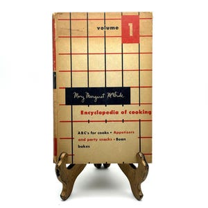 May include: Vintage cookbook, "Encyclopedia of Cooking, Volume 1" by Mary Margaret McBride, displayed on a decorative gold-toned stand. The book cover is tan with a red and black grid design, and the title is prominently displayed.
