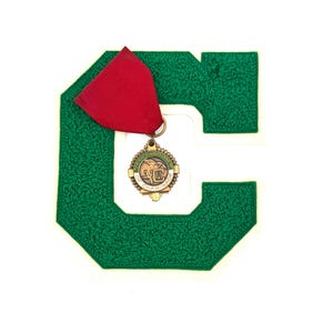 May include: A green felt letter "C" with a white border, featuring a gold-toned medal with a red ribbon. The medal has text and a crest design. The letter is textured and stands out against the white background.