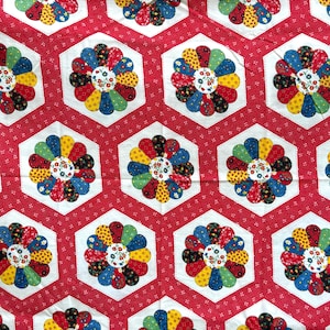 May include: A red fabric with a repeating pattern of colorful flower-like designs. Each flower is composed of petal shapes in various colors, arranged within a white hexagon. The background is red with small white dots.