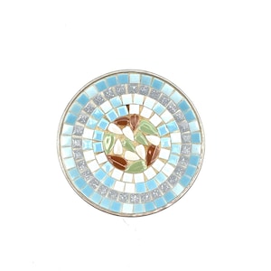 May include: A round mosaic tabletop with a light blue, silver, brown, and green design. The mosaic is made of small, square tiles arranged in concentric circles. The outer ring is light blue, followed by silver, white, brown, and green tiles.