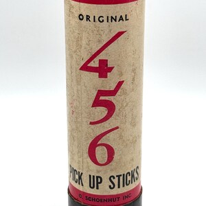 May include: A vintage "Original 456 Pick Up Sticks" game in a cylindrical cardboard container. The container has a tan background with red text and numbers. The top and bottom are black.