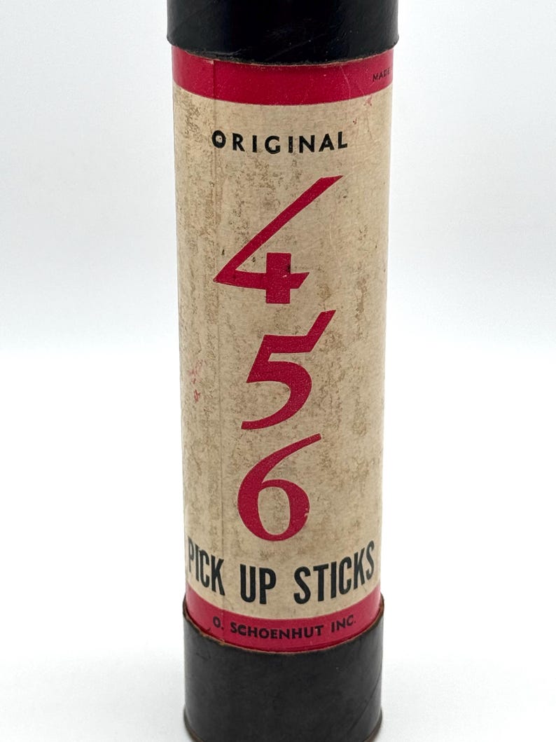 May include: A vintage "Original 456 Pick Up Sticks" game in a cylindrical cardboard container. The container has a tan background with red text and numbers. The top and bottom are black.