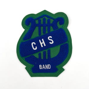 May include: A green and blue patch with the text "CHS BAND". The patch has a shield shape with a blue lyre design and white lettering. The patch is made of felt.