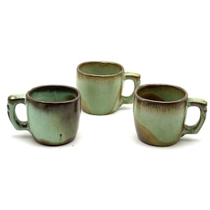 May include: Three ceramic mugs featuring a reactive glaze finish in shades of sage green and dark brown. Each mug has a rounded body, a sturdy side handle, and a smooth rim. The glaze displays a dripped effect with darker tones near the top.