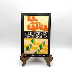 May include: A vintage book titled "SILK SCREEN TECHNIQUES" with a black cover and colorful geometric designs. The book is displayed on a decorative gold-colored stand against a white background. The author's name, BIEGELEISEN AND COHN, is printed at the bottom.