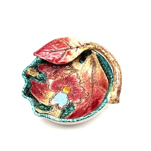 May include: A ceramic decorative bowl with a floral design. The bowl features a red flower with a light blue center and red and yellow leaves. The rim is turquoise and the handle is brown.