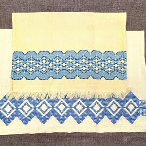 May include: Two cream-colored embroidered towels. The top towel has a blue and white geometric design across the center. The bottom towel has a similar design at the bottom edge. Both towels are made of a woven fabric.
