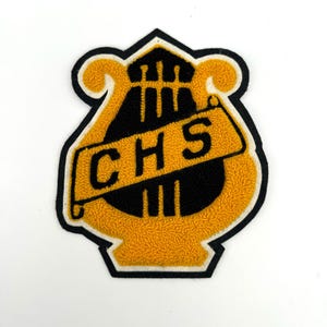 May include: A yellow and black patch with the letters "CHS" on a banner. The patch is shaped like a lyre and has a black border. The background is white.