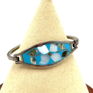 May include: Silver-tone bracelet with a turquoise center featuring a flower design. The flower is made of white and iridescent shell pieces. The bracelet is displayed on a beige cone-shaped stand.
