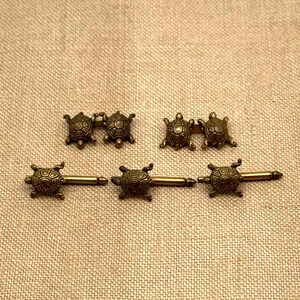 May include: Six antique bronze-colored turtle-shaped jewelry pieces. Two pairs of turtle cufflinks and three turtle-shaped tie tacks are displayed on a tan fabric surface. The turtles have detailed shell patterns.