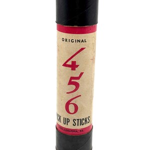 May include: A vintage cylindrical container for "Pick Up Sticks" with a tan body, black top and bottom, and a red band. The text "ORIGINAL" and the numbers "4 5 6" are in red. The bottom reads "PHILADELPHIA, PA."