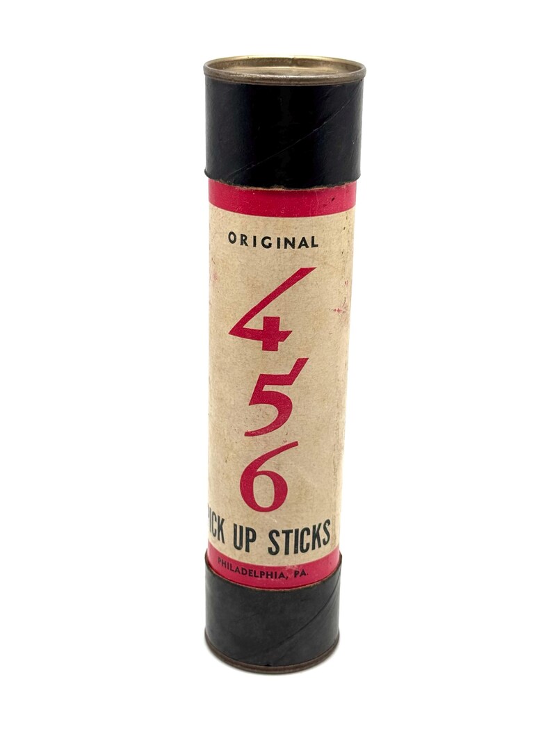 May include: A vintage cylindrical container for "Pick Up Sticks" with a tan body, black top and bottom, and a red band. The text "ORIGINAL" and the numbers "4 5 6" are in red. The bottom reads "PHILADELPHIA, PA."