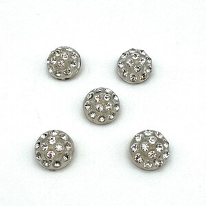 May include: Five silver-toned, round buttons with a domed surface. Each button is encrusted with small, clear rhinestones, creating a sparkling effect. The buttons are evenly spaced against a white background, suitable for sewing or craft projects.