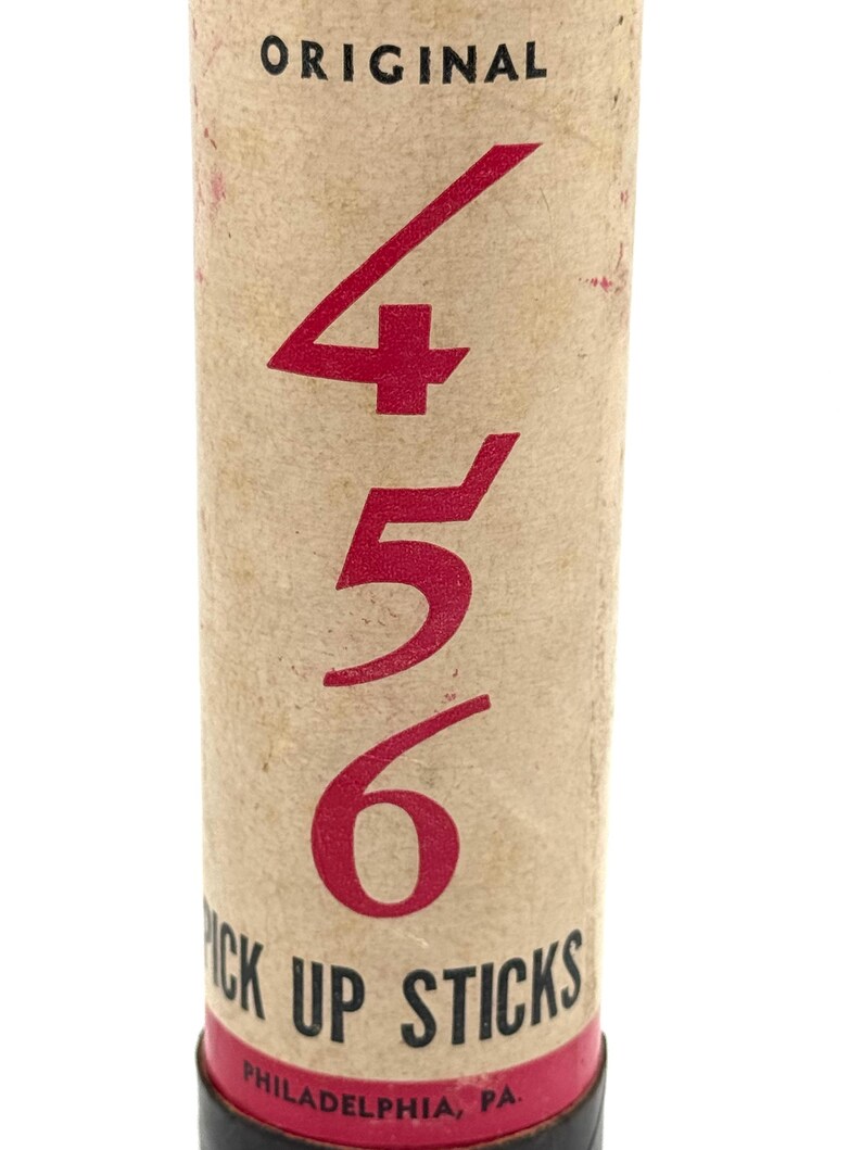 May include: A vintage tube of "Pick Up Sticks" game. The cylindrical container is tan with red text and numbers. The numbers "456" are prominently displayed. The text "ORIGINAL" is at the top, and "PICK UP STICKS" and "PHILADELPHIA, PA" are at the bottom.