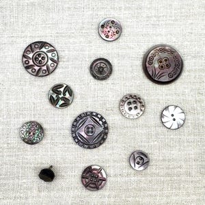 May include: A collection of various iridescent buttons with intricate designs, including floral, geometric, and circular patterns. The buttons are in shades of gray, black, and white, and are arranged on a light beige fabric.
