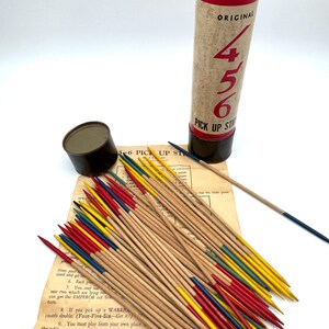 May include: A vintage 4-5-6 Pick Up Sticks game set. The game includes a cylindrical container with red and black accents, a small metal container, a rule sheet, and a collection of wooden sticks with colored tips.