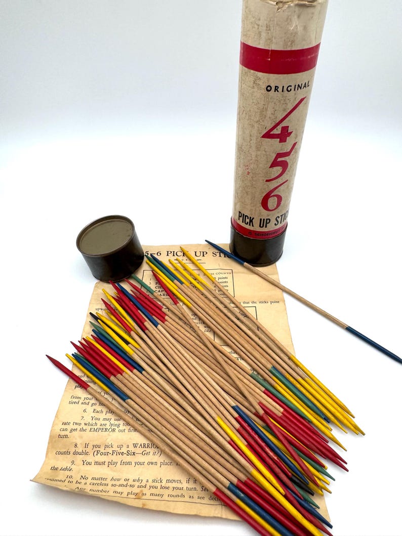 May include: A vintage 4-5-6 Pick Up Sticks game set. The game includes a cylindrical container with red and black accents, a small metal container, a rule sheet, and a collection of wooden sticks with colored tips.