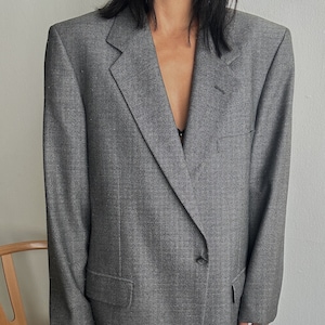 Puede incluir: Una chaqueta gris con un cierre de un botón. La chaqueta tiene un corte largo y relajado y una silueta ligeramente oversized.