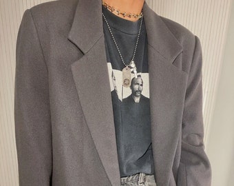 Könnte beinhalten: Ein grauer Blazer mit einem einzigen Knopfverschluss. Der Blazer hat einen Schalkragen und eine Tasche auf der linken Seite. Der Blazer wird über einem schwarzen T-Shirt mit einem grafischen Aufdruck und einer silbernen Kettenkette getragen.