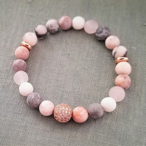 May include: A pink, gray, and white beaded bracelet with a rose gold charm. The bracelet has a rose gold accent bead with a clear crystal center.