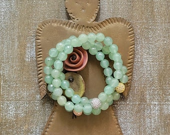 May include: A double strand of light green gemstone beads with gold and silver accents. The beads are arranged in a circle on a rustic brown background.