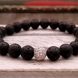 Puede incluir: Pulsera de cuentas de piedra de lava negra con una sola cuenta plateada brillante. La pulsera está ensartada en una banda elástica. Las cuentas son redondas y tienen una textura porosa. Se muestra sobre una superficie de madera clara.