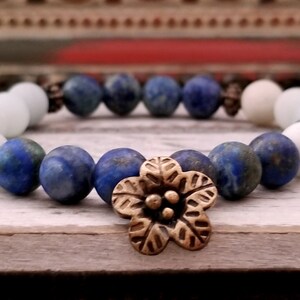 以下が含まれることがあります： 青と白のビーズブレスレットに、ブロンズのフラワーチャームが付いています。ブレスレットは、ラピスラズリとアクアマリンのビーズで作られています。