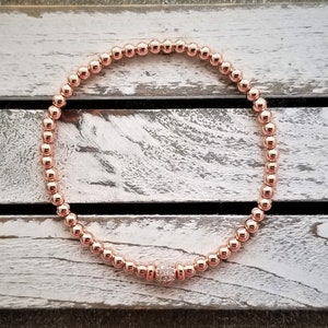 May include: A rose gold beaded bracelet with a single larger rose gold bead accented with small clear stones.