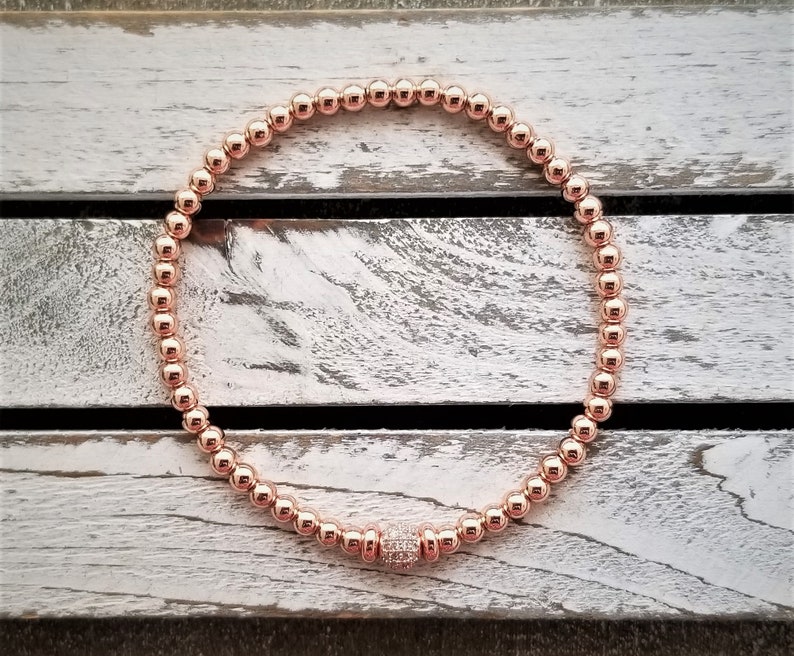May include: A rose gold beaded bracelet with a single larger rose gold bead accented with small clear stones.