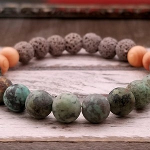 Puede incluir: Un brazalete hecho de turquesa, roca volcánica y cuentas de madera. El brazalete es una mezcla de colores verde, marrón y naranja.