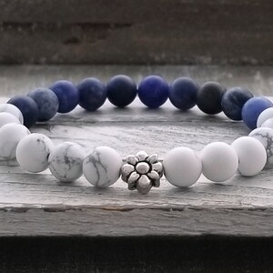 May include: A white and blue beaded bracelet with a silver flower charm. The bracelet is made of white howlite and blue lapis lazuli beads.