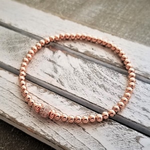 May include: A rose gold beaded bracelet with a single larger bead accented with small clear stones.