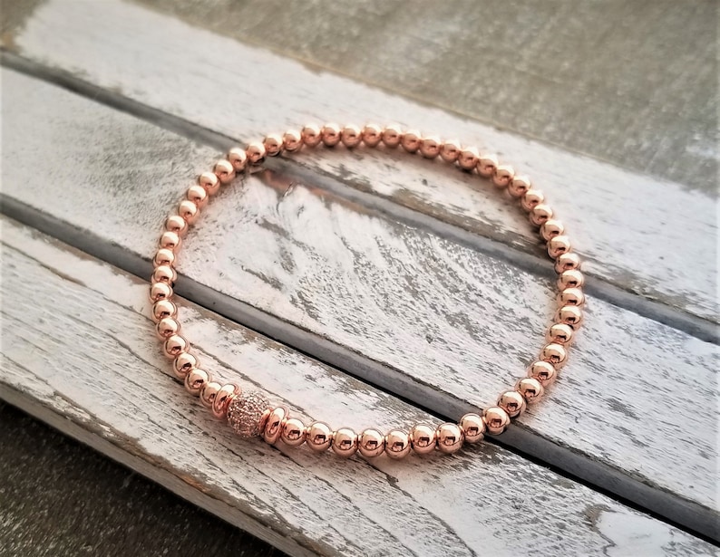 May include: A rose gold beaded bracelet with a single larger bead accented with small clear stones.