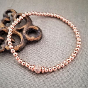 May include: A rose gold beaded bracelet with a single larger bead in the center that is encrusted with smaller stones.