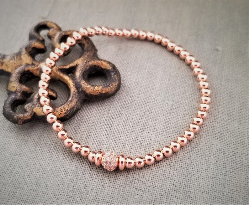 May include: A rose gold beaded bracelet with a single larger bead in the center that is encrusted with smaller stones.