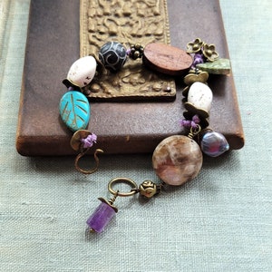 Op de afbeelding: Een veelkleurige armband met verschillende kralen, waaronder een grote, bruine, ovale kraal, een kleinere, zwarte kraal met een wit ontwerp, een turquoise bladvormige kraal en een paarse amethistkraal. De armband is geregen met paarse en turquoise draad.