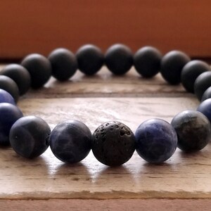 Puede incluir: Un brazalete de cuentas con cuentas negras, azules y gris oscuro. El brazalete tiene una sola cuenta gris oscuro en el centro.