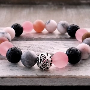 Puede incluir: Un brazalete hecho con cuentas rosas, grises y negras, con un dije de plata en el centro.