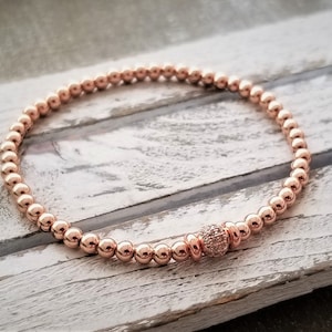 May include: A rose gold beaded bracelet with a single larger bead in the center that is encrusted with smaller clear stones.