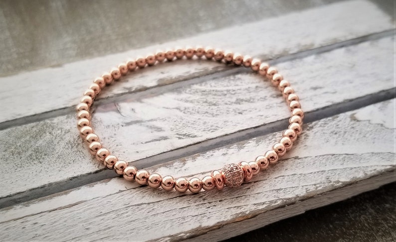 May include: A rose gold beaded bracelet with a single larger bead in the center that is encrusted with smaller clear stones.