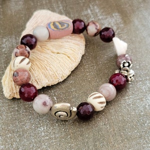 Op de afbeelding: Een armband gemaakt met rode, witte en bruine kralen, met zilveren accenten. De armband ligt op een lichtbruin oppervlak.